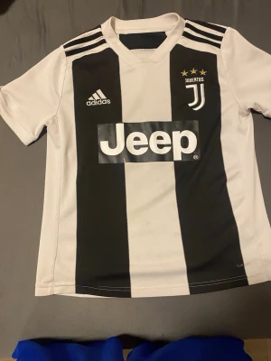 Juventus matchtröja Adidas svartvit - Juventus officiella matchtröja från Adidas i svartvitt med klassiska breda ränder. Tröjan har korta ärmar, klubbmärke och tre stjärnor på bröstet samt Jeep-logga framtill. Tillverkad i lätt och ventilerande material, perfekt för fotbollsträning eller match.