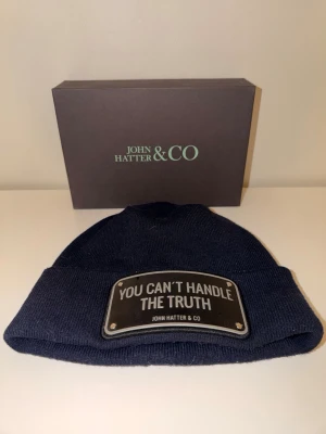John hatter & co navy blå mössa - Det här är en marinblå mössa från John Hatter & Co. Mössan har en metallplatta framtill med texten ”YOU CAN’T HANDLE THE TRUTH”. Produkten visas tillsammans med sin märkesförsedda ask i bakgrunden.