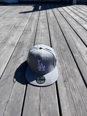 Ny Grå LA-keps från New Era - Helt ny fitted grå keps med LA-logga framtill och klassisk rak skärm. Kepsen är från New Era och modellen är 59FIFTY. Perfekt för dig som gillar streetwear och vill ha en stilren accessoar till din outfit. Storlek 55,8 cm