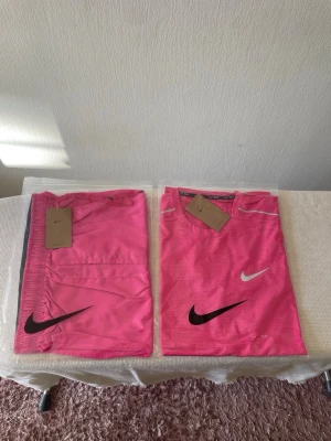 Rosa träningsset från Nike - Säljer ett rosa träningsset från Nike med t-shirt och shorts. Båda plaggen har tydliga Nike-loggor i svart och vitt. Materialet är lätt och andas, perfekt för träning eller löpning. Snygg sportig design med diskreta ränder på t-shirten.
