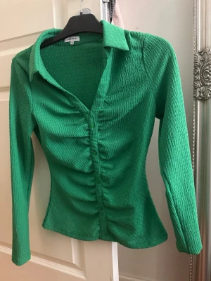 Grön långärmad skjorta med rynk - Snygg grön skjorta med lång ärm och rynkdetaljer längs knäppningen. Skjortan har klassisk krage och figurnära passform, perfekt för en trendig look. Materialet är lätt och stretchigt för extra komfort.
