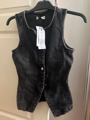 Svart jeansväst med knappar - Snygg svart jeansväst i figurnära modell med knäppning framtill. Västen har markerade sömmar och en klassisk rund halsringning. Perfekt att styla över en t-shirt eller skjorta för en cool och trendig look.