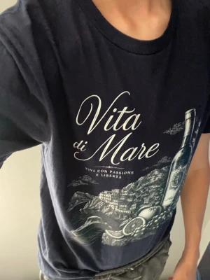 Mörkblå t-shirt med tryck Vita di Mare - Snygg mörkblå t-shirt med stort vitt tryck på framsidan där det står 'Vita di Mare' och motiv av en vinflaska, hav och landskap. T-shirten har rund hals och korta ärmar. Perfekt för dig som gillar stilrena och coola prints.