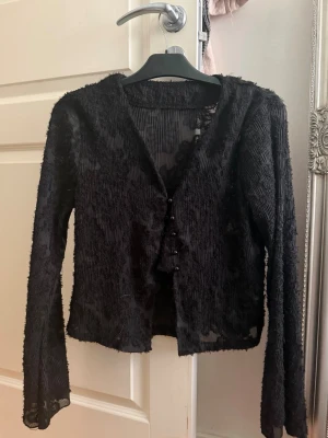 Svart transparent kofta med knappar - Säljer en svart, transparent kofta med lång ärm och dekorativa knappar framtill. Materialet är ribbat och har en lätt glansig finish som ger ett coolt och trendigt intryck. Perfekt att styla över ett linne eller topp för en extra touch.