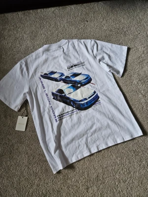 Vit t-shirt Aimé Leon Dore med biltryck - Vit t-shirt från Aimé Leon Dore med tryck av blå sportbilar på ryggen och liten logga framtill. Klassisk passform med rund hals och korta ärmar. Perfekt för dig som gillar streetwear och snygga prints.