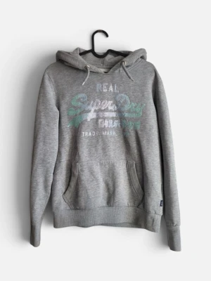 Grå hoodie med tryck från Superdry - Säljer en grå hoodie från Superdry med stort tryck i vitt och turkost på bröstet. Hoodien har huva med dragsko, magficka och ribbade muddar vid ärmar och nederkant. Perfekt för en avslappnad och cool stil. Kan vara lite smutsig vid ärmarna men igen större defekter.