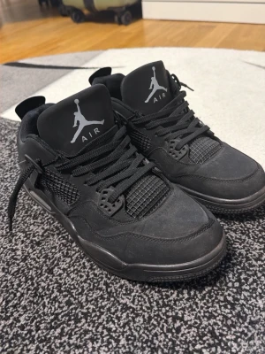 Svarta Air Jordan 4 sneakers - Säljer ett par helsvarta Air Jordan 4 sneakers med klassisk Jumpman-logga på plösen. Skorna har meshdetaljer på sidorna och tåboxen, samt robusta snören och en chunky sula. Materialet är en mix av mocka och textil för en cool streetlook.