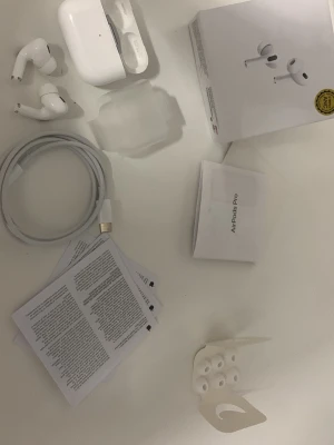Apple AirPods Pro Unboxing - These images show the contents of a new Apple AirPods Pro box. The first image displays all items laid out: the AirPods Pro earbuds, charging case, charging cable, user manuals, and extra ear tips. The second image gives a closer look at the earbuds and charging case.