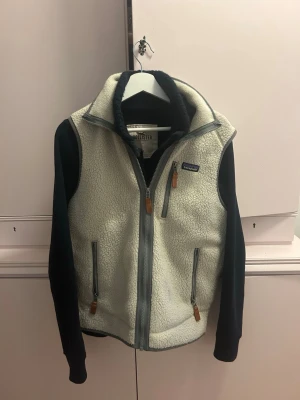 Vit fleeceväst från Patagonia S - Säljer en snygg vit fleeceväst från Patagonia i storlek S. Västen har dragkedja framtill, tre fickor med dragkedja och detaljer i läder. Perfekt för lager-på-lager och outdoor-vibbar. Klassisk Patagonia-logga på bröstet.