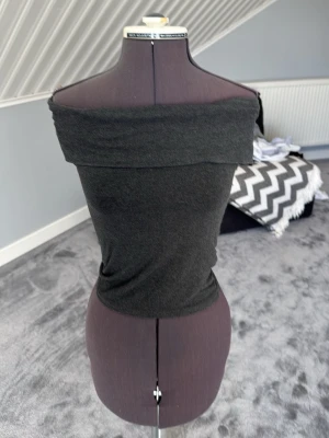 Mörkgrå offshoulder topp - Säljer en stilren mörkgrå offshoulder topp med tight passform. Toppen är ärmlös och har en bred kant upptill som ger en snygg siluett. Perfekt att matcha med jeans eller kjol för en trendig look.