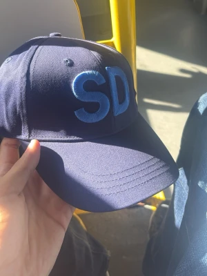 Mörkblå keps med SD-brodyr - Säljer en mörkblå keps med stora broderade bokstäver 'SD' i ljusblått framtill. På sidan finns en liten broderad bi tillsammans med texten 'SD'. Justerbar rem baktill för perfekt passform. Klassisk böjd skärm och ventilerande hål upptill.