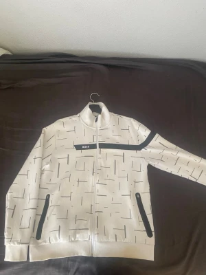 Vit tröja med svart mönster från BOSS - Snygg vit hoodie från BOSS med svart grafiskt mönster och svarta detaljer på dragkedjor och bröstet. Dragkedja framtill och två fickor med svarta dragkedjor. Hög krage och modern passform, perfekt för en trendig streetwear-look.
