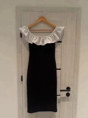 Off shoulder Svart Klänning med Vit Volang - Säljer denna jättefina figurnära klänning från H&M. Den perfekta blandningen av sofistikerat och feminint med sin vackra off-shoulder-volang i kontrastfärg!  • Märke: H&M. • Modell: Figurnära (bodycon) med en stor, vit volang upptill. • Passform: Stretchigt och bekvämt material som följer kroppen (95% polyester, 5% elastan). • Storlek: S (EU 36).  • 🏠 Rök- och djurfritt hem. • ⚡ Snabb leverans: Jag paketerar och skickar inom 24 timmar!  