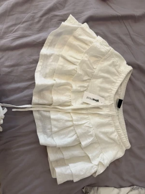 Vita volangshorts från Gina Tricot - Supersöta vita shorts från Gina Tricot med flera volanglager och snörning i midjan. De har en elastisk midja och är perfekta för varma dagar. Luftigt material och trendig design som ger en romantisk vibe.