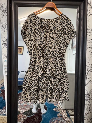 Leopardmönstrad volangklänning - Kort klänning med leopardmönster i svart och beige. Klänningen har volanger och en lös passform med rund halsringning och korta ärmar. Perfekt för dig som vill sticka ut med ett trendigt djurmönster.