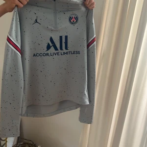 Grå långärmad träningströja PSG x Jordan - Säljer en grå långärmad träningströja från PSG och Jordan med blå logga och text, samt röda och blå ränder på ärmarna. Tröjan har halv dragkedja och stänkmönster över hela. Perfekt för fotbollsträning eller sport. Xl junior .