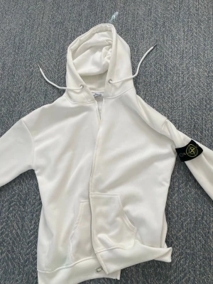 Vit hoodie från Stone Island - Säljer en clean vit hoodie från Stone Island med klassisk huva och dragsnören. Hoodien har en ficka framtill och den ikoniska Stone Island-loggan på ärmen. Perfekt för dig som gillar streetwear och vill ha något stilrent i garderoben.