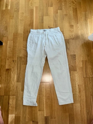 Vita linnebyxor från ZARA - Säljer ett par vita linnebyxor från ZARA med avslappnad passform och dragsko i midjan. Byxorna har resår i midjan och sidofickor, perfekta för en chill och stilren look. Materialet är lätt och luftigt linne, vilket gör dem sköna att bära under varma dagar.