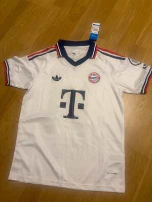 Olise bayern 26/27 borta S - Snygg vit FC Bayern München matchtröja från Adidas med mörkblå och röda detaljer på krage och ärmslut. Stort T-Mobile-tryck på bröstet, klubbmärke och Adidas-logga. Nummer 17 och namnet Olise på ryggen. Tillverkad i lätt och ventilerande material.