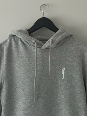 Grå hoodie med vit logga ifrån rs  - Snygg grå hoodie med huva och dragsnören samt en diskret vit logga på bröstet. Hoodien har en klassisk känguruficka framtill och är tillverkad i mjukt material som känns skönt mot huden. Perfekt för en avslappnad och stilren look.