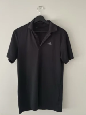 Svart pikétröja från Adidas använd fåtal gånger - Klassisk svart pikétröja från Adidas med korta ärmar och diskret logga på bröstet. Tröjan har en enkel krage och knappslå framtill. Perfekt för en sportig och stilren look.
