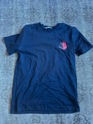 Moncler t-shirt  - Säljer en mörkblå t-shirt med korta ärmar och en liten patch med rött och vitt tryck på bröstet. T-shirten har en klassisk passform och rund hals. Perfekt till jeans eller shorts för en enkel och snygg look.