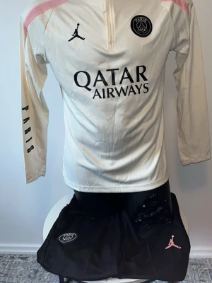 PSG träningsset i vitt och svart - Säljer ett PSG träningsset med långärmad vit tröja och svarta byxor. Tröjan har rosa detaljer, PSG-logga och Jordan-logga samt Qatar Airways-tryck. Byxorna är svarta med rosa detaljer, PSG-logga och texten PARIS längs benet. Perfekt för fotbollsträning eller chill.