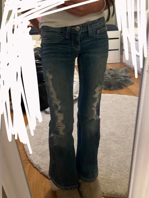 Bootcut jeans - Säljer ett par blå bootcut jeans med hål på framsidan av båda benen. Jeansen har klassisk femficksmodell, låg midja och är gjorda i denim. Perfekta för en avslappnad och trendig look.