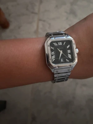 Fin klocka  - Stilren klocka i silverfärgat metallarmband och fyrkantigt boett. Svart urtavla med romerska siffror och datumvisning. Klassisk design som passar till många olika stilar.