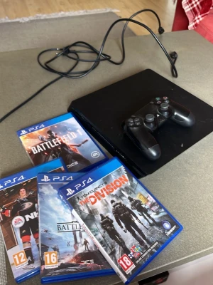 PlayStation 4 Slim med 4 spel  - Säljer en svart PlayStation 4 Slim med tillhörande DualShock 4-handkontroll och Battlefield, Nhl23, starwars battlefront, och the division . Konsolen har en stilren, platt design och är tillverkad i plast. Perfekt för gaming och filmkvällar med vänner.