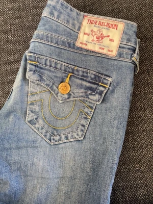 True Religion ljusblå utsvängda jeans - Säljer ett par ljusblå jeans från True Religion med breda utsvängda ben och slitna detaljer. Jeansen har klassiska kontrastsömmar, stora fickor med lock och guldfärgade knappar. Perfekta för dig som gillar en retro och avslappnad stil.