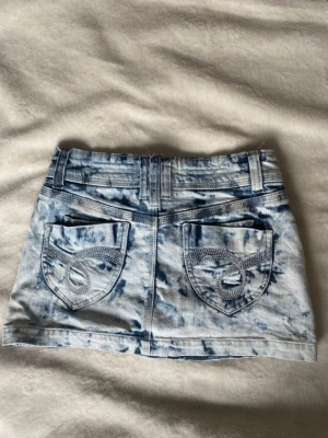 Blåvit kort jeanskjol med mönster - Säljer en snygg blåvit kort jeanskjol med batikmönster och dekorativa bakfickor med detaljer. Kjolen har klassiska fickor fram och bak, bälteshällor och knappstängning. Perfekt för dig som vill ha en trendig och unik denimlook. Storlek 6!