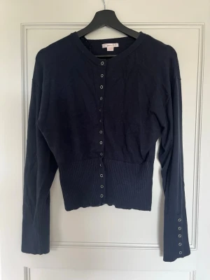 Marinblå kofta från Gina Tricot - Stilren marinblå kofta från Gina Tricot med långa ärmar och ribbade muddar. Knäppning framtill med silverfärgade tryckknappar och extra detaljer vid ärmsluten. Perfekt att slänga över en topp för en enkel men snygg look.