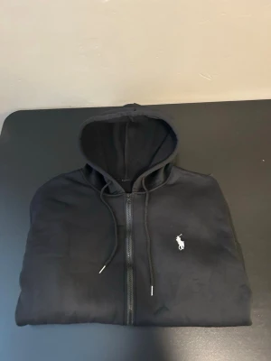 Svart zip hoodie från Polo Ralph Lauren - Snygg svart hoodie med dragkedja från Polo Ralph Lauren. Klassisk design med huva och justerbara snören, samt den ikoniska vita logon broderad på bröstet. Perfekt för en chill och stilren look.