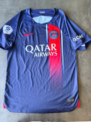 PSG matchtröja blå/röd Nike - Snygg Paris Saint-Germain matchtröja i blått med röd vertikal rand och vita detaljer. Tröjan har PSG-logga, Qatar Airways-tryck och Nike swoosh på bröstet. Tillverkad i lätt och ventilerande material, använd ett par ggr så i väldigt bra skick. 
