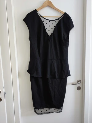 Svart långklänning - Stilren svart långklänning med v-ringning och transparent mesh upptill samt nertill, dekorerad med svarta prickar. Klänningen har en figurnära passform och korta ärmar. Perfekt för dig som vill ha en elegant look med en twist.