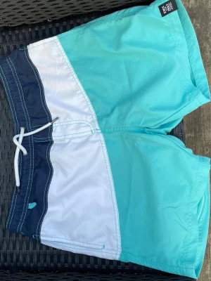 Turkosa och vita badshorts - Säljer ett par snygga badshorts i turkos, vitt och marinblått. De har dragsko i midjan och en bakficka med kardborre. Klassisk färgblockad design och tillverkade i snabbtorkande material, perfekta för stranden eller poolen.