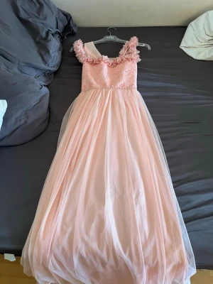 Bröllops klänning  - Säljer en drömmig ljusrosa långklänning med volangdetaljer vid axlarna och ett fluffigt, svepande kjolparti i flera lager. Klänningen har ärmlös design och vacker spets upptill. Perfekt för speciella tillfällen! Storlek 152, 12-13 år! 