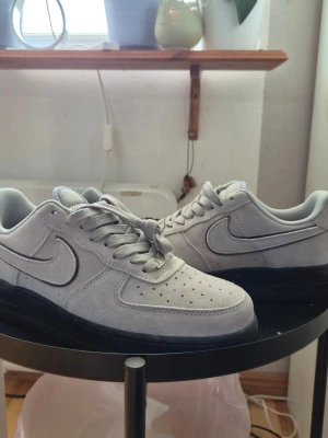 Nike Air Force 1 låga sneakers grå - Snygga Nike Air Force 1 sneakers i ljusgrå mocka med klassisk svart sula och vit logga på plösen. Skorna har perforerad tå, vita skosnören och den ikoniska Swoosh-loggan på sidan. Perfekt för dig som gillar stilrena och tidlösa sneakers.