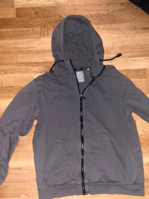 Grå hoodie med dragkedja från Etirel - Säljer en grå hoodie från Etirel med dragkedja framtill och två stora fickor. Tröjan har huva med dragsko och ribbade muddar vid ärmslut och nederkant. Perfekt för chill dagar eller när du vill ha en enkel och stilren look.
