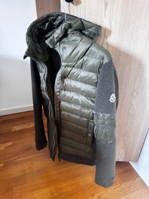 Grön hybrid cardiganjacka Moncler. Tar även byten  - Snygg grön cardiganjacka från Moncler med quiltad front och huva i blankt material samt stickade ärmar och rygg. Jackan har dragkedja, fickor med knapp och Moncler-logga på ärmen. Perfekt för lager-på-lager och streetstyle.med qr kod och tagg i armen 