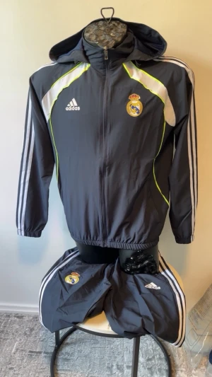 Adidas Real Madrid träningsset - Mörkgrått träningsset från Adidas med Real Madrid-logga på både jacka och byxor. Setet har vita och neongula detaljer, dragkedja framtill och huva på jackan. Klassiska Adidas-ränder längs ärmar och ben. Perfekt för fotbollsträning eller chill.
