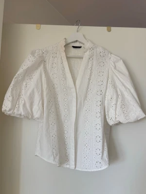 Vit blus med puffärm och broderie från Zara - Söt vit blus med puffiga trekvartsärmar och vackert broderie anglaise-mönster. Blusen har v-ringning och är gjord i ett lätt bomullstyg som känns luftigt och somrigt. Perfekt till jeans eller kjol för en trendig look.