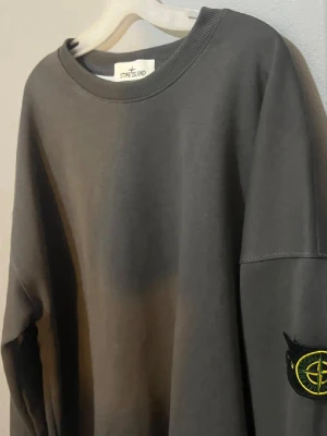 Grå sweatshirt från Stone Island - Säljer en stilren grå sweatshirt från Stone Island med klassisk rund halsringning och ribbade muddar. Tröjan har den ikoniska Stone Island-loggan på ärmen och är tillverkad i mjukt bomullsmaterial. Perfekt för en avslappnad och trendig look. Tröjan är L men är lite större i storlek 