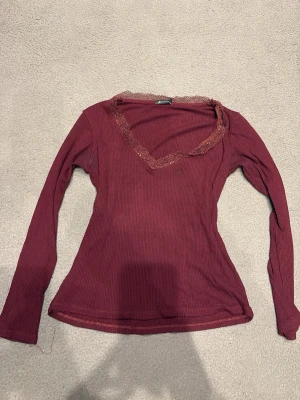Vinröd ribbad topp från Stradivarius - Vinröd långärmad topp från Stradivarius med ribbad struktur och spetsdetalj längs v-ringningen. Figurnära passform som ger en snygg siluett. Perfekt att styla med jeans eller kjol för en trendig look.