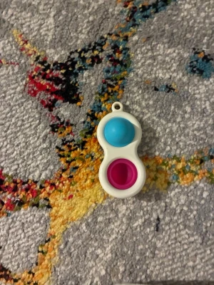 Fidget popper i blått och rosa - En liten fidget popper i plast med två bubblor, en blå och en rosa. Perfekt att ha på nyckelringen eller i fickan för att hålla händerna sysselsatta. Vit ram i åttaformad design och hål för att fästa i nyckelknippa.
