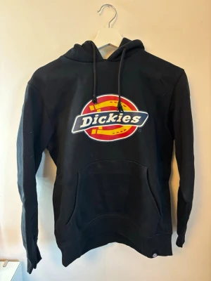 Svart Dickies hoodie  - Svart hoodie från Dickies med stor färgglad logga på bröstet. Klassisk modell med huva, dragsnören och känguruficka framtill. Mjuk och bekväm bomullsblandning.