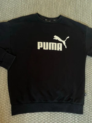 Svart sweatshirt från Puma - Svart sweatshirt från Puma med klassisk vit logga och text på bröstet. Tröjan har rund halsringning, ribbade muddar vid ärmslut. 