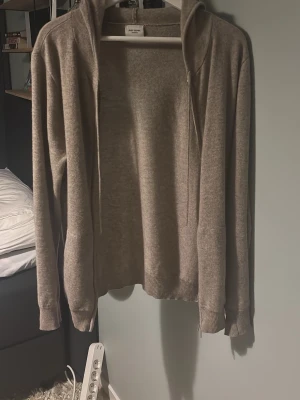 John Henric zip  - Mysig beige hoodie i mjukt stickat material med dragkedja framtill och ribbade muddar vid ärmar och nederkant. Klassisk huva och enkel design utan tryck eller detaljer. Perfekt för en avslappnad stil.