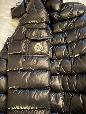 Svart dunjacka från Moncler - Säljer en svart, glansig dunjacka från Moncler med klassisk logotyp på ärmen och praktisk ficka med lock. Jackan har breda kanaler och hög krage, perfekt för kalla vinterdagar. Stilsäker och varm med exklusiv känsla.